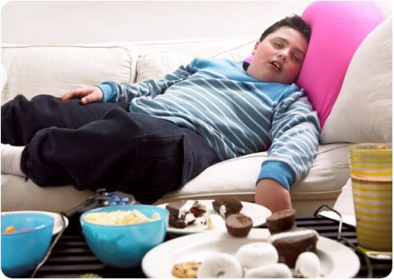 Un garcon en surpoids endormi devant des plats de chips et de gâteaux